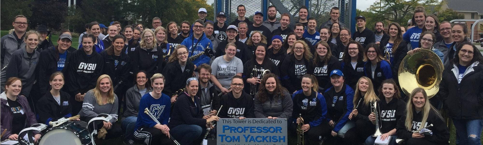 The Laker Alumni Marching Band posing in front of the LMB tower, presenting a dedication plaque that reads "This tower is dedicated to Professor Tom Yackish"